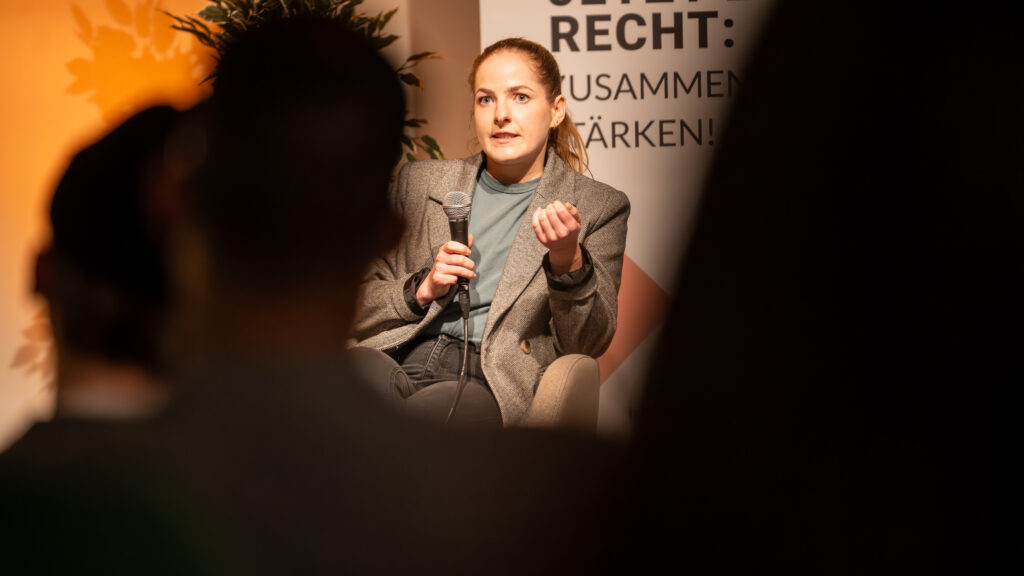 Frau sitzt mit Mikrofon vor Banner und spricht in Publikum, im Vordergrund unscharfe Silhouetten von Zuhörern
