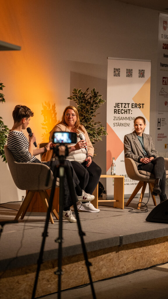 Drei Frauen sitzen auf Stühlen auf einer Bühne, eine spricht in ein Mikrofon, davor steht eine Kamera auf einem Stativ, im Hintergrund ein Banner mit Text und QR-Codes.
