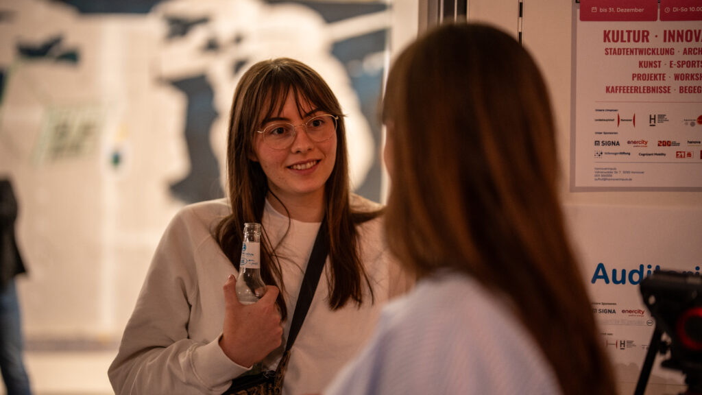 Junge Frau mit Brille und langem Haar hält eine Flasche und spricht mit einer anderen Person, die nur von hinten zu sehen ist