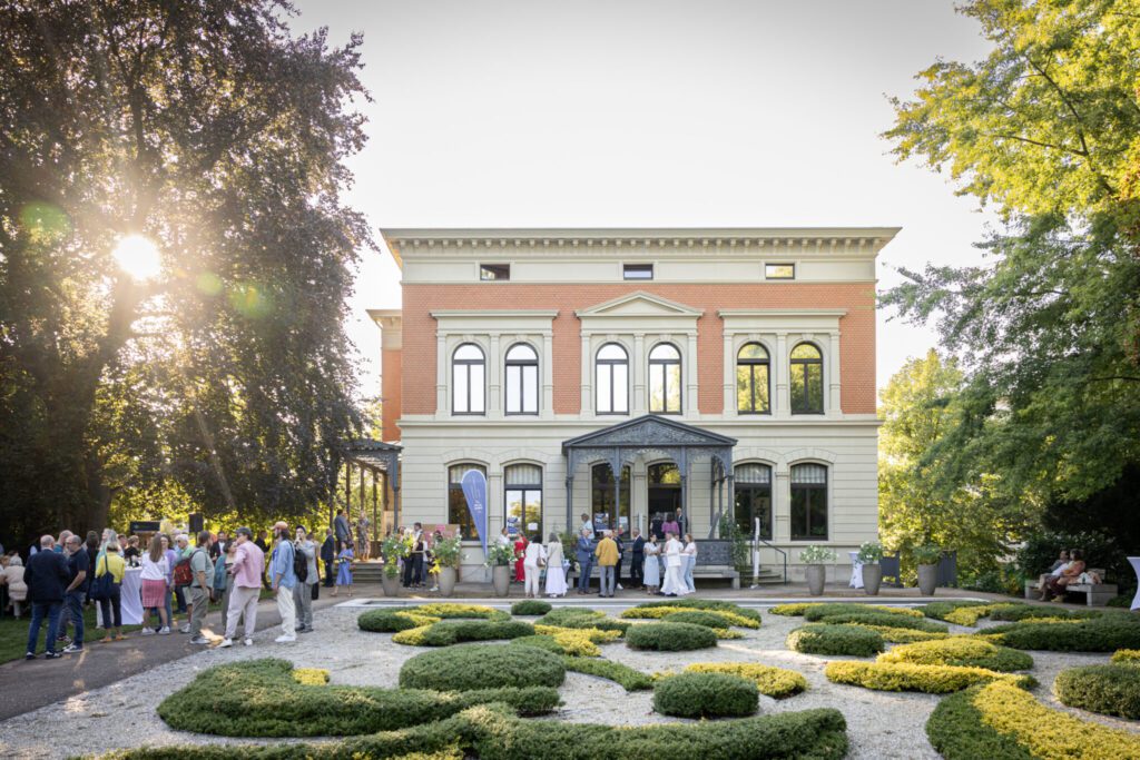 Menschenmenge vor einem zweistöckigen historischen Gebäude mit symmetrischer Fensteranordnung und dekorativem Vorgarten bei Sonnenuntergang