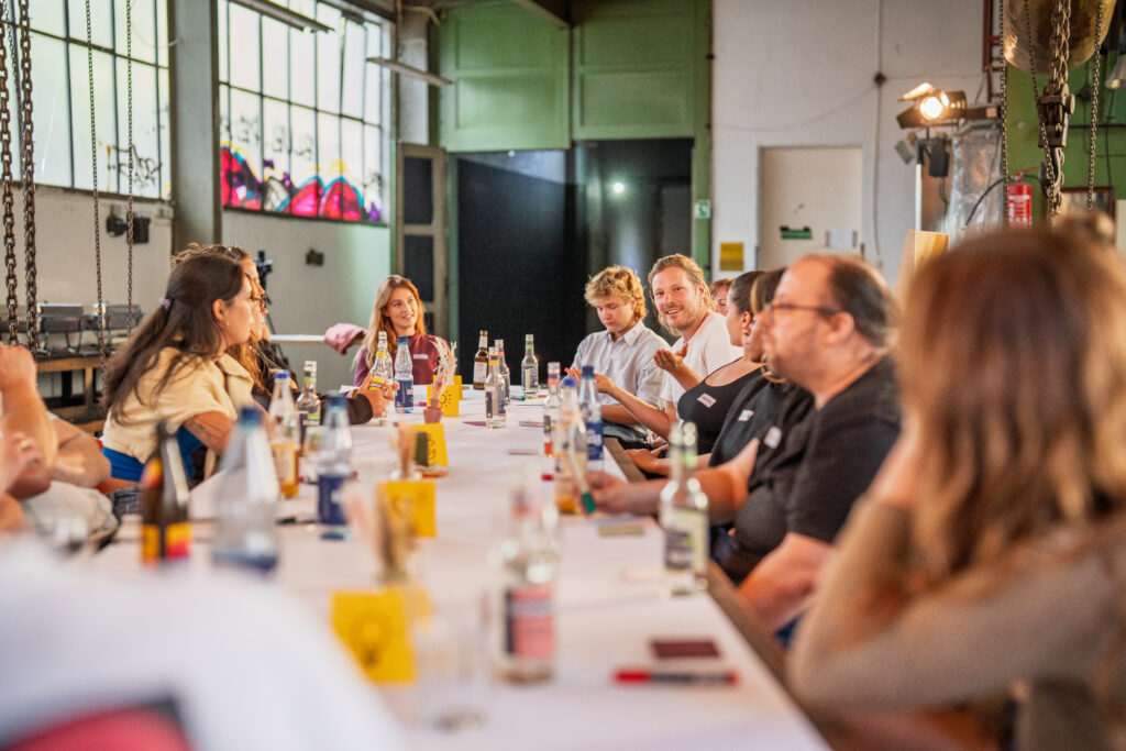 Menschen sitzen an einem langen Tisch in einem hellen Raum und unterhalten sich