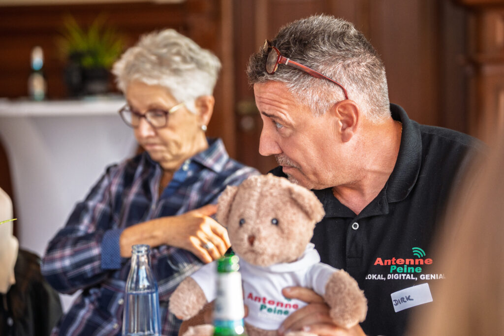 Mann mit grauem Haar und Brille auf dem Kopf hält Teddybär mit weißem T-Shirt und sitzt neben älterer Frau mit kurzen grauen Haaren und Brille in einem Raum mit Holzmöbeln