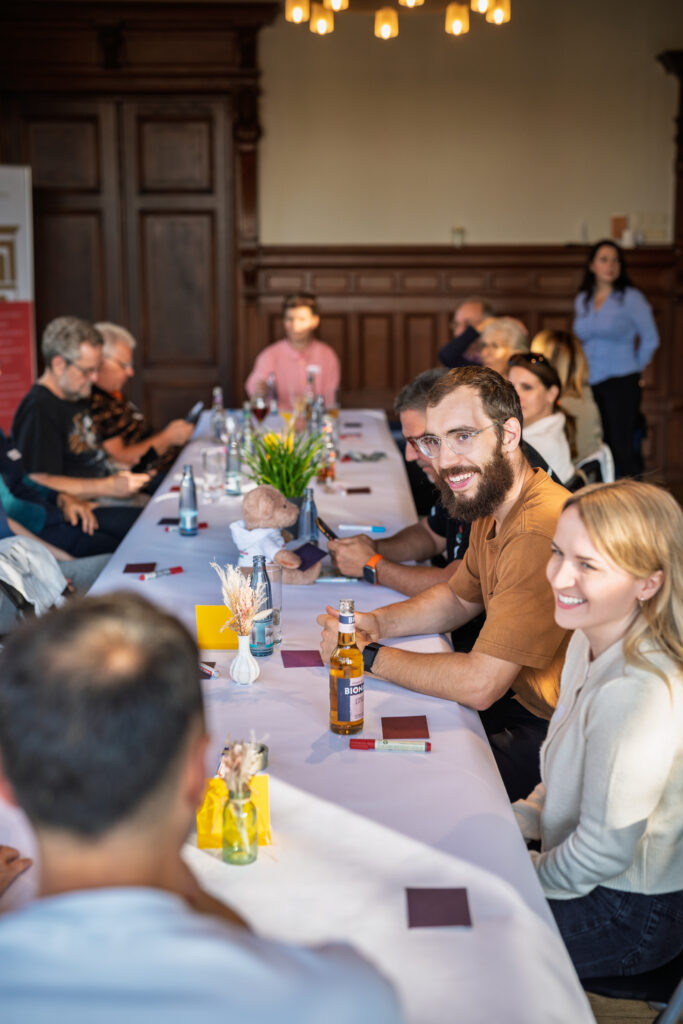 Gruppe von Menschen sitzt an einem langen Tisch in einem Raum mit Holzvertäfelung, zwei Personen im Vordergrund lächeln in die Kamera