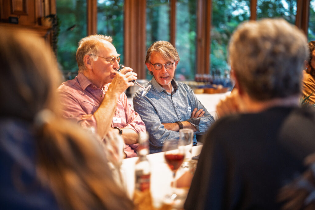 Zwei ältere Männer sitzen an einem Tisch und unterhalten sich, einer trinkt aus einem Glas