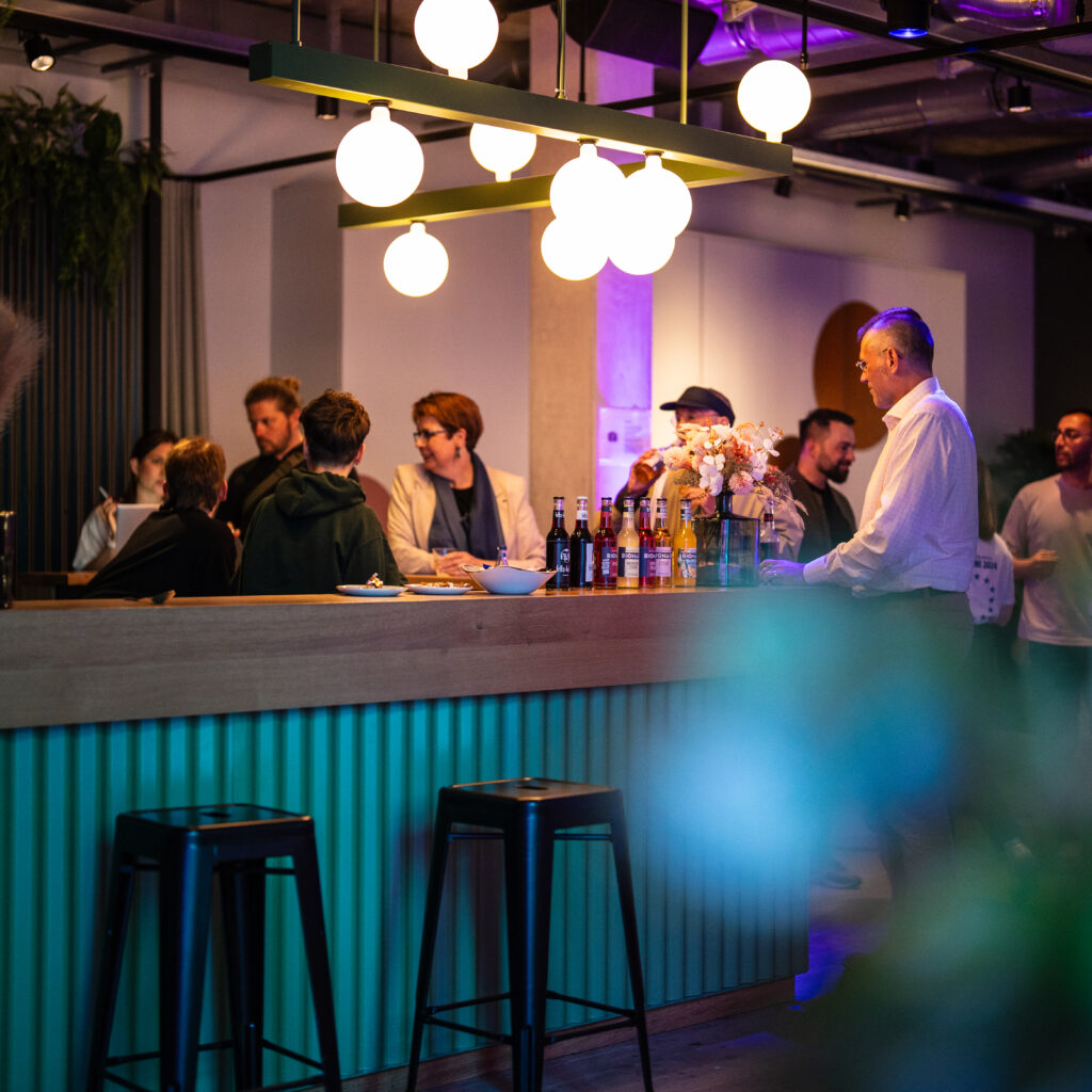 Mehrere Personen unterhalten sich an einer Bar mit hängenden Kugelleuchten und einer Theke mit Flaschen und Blumen in einem modernen Innenraum