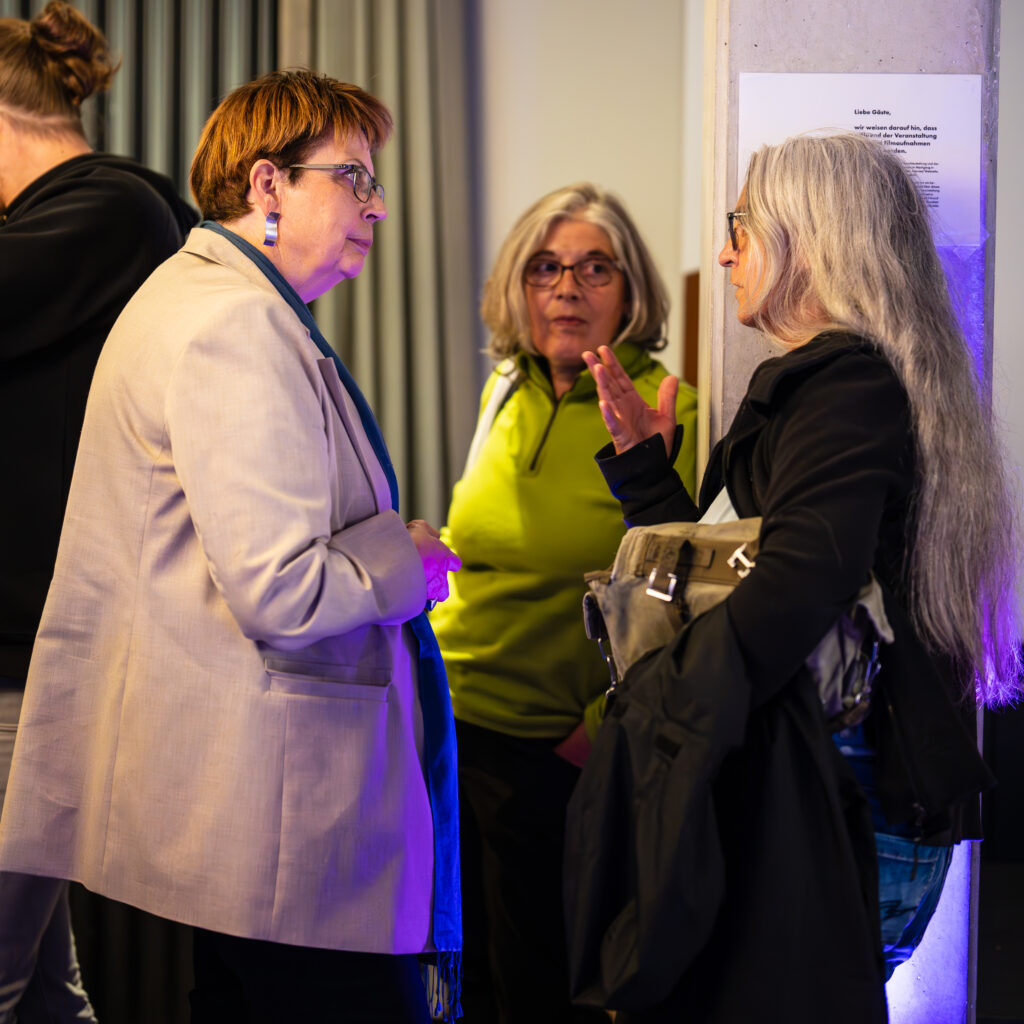 Drei Frauen im Gespräch vor einer Wand mit einem Aushang, eine trägt eine beige Jacke, eine eine grüne Jacke, die dritte eine schwarze Jacke und hält eine Tasche.