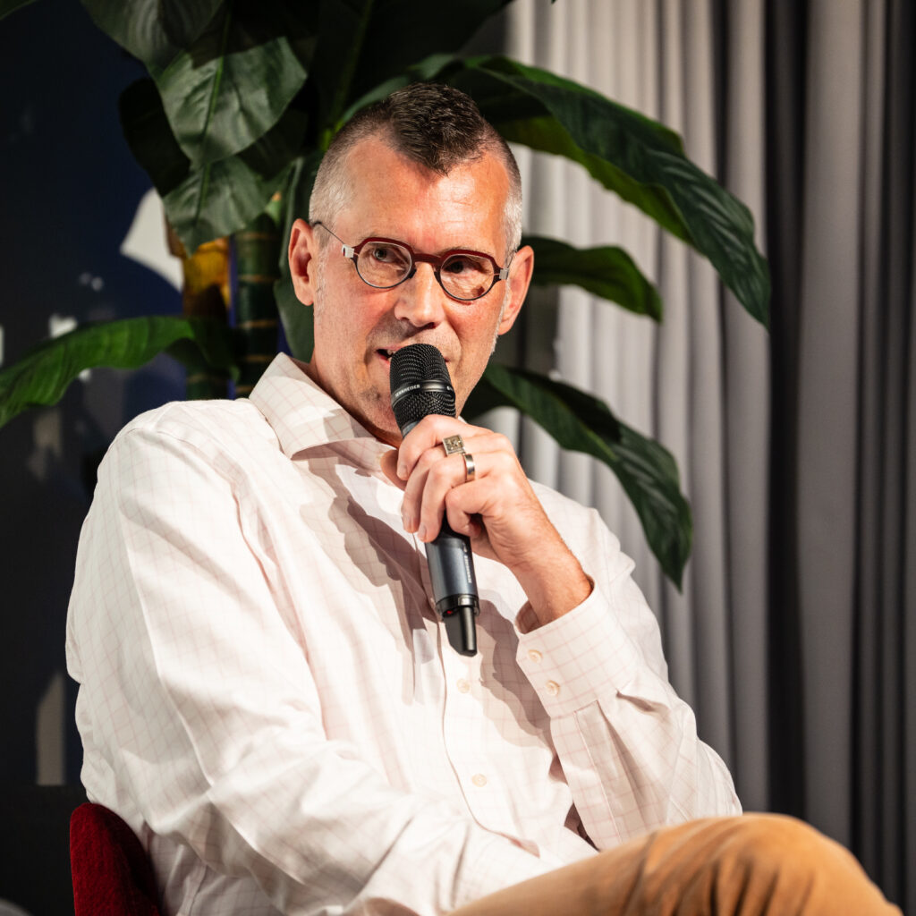 Mann mit Brille und weißem Hemd spricht in ein Mikrofon vor dunklem Hintergrund mit großen Blättern