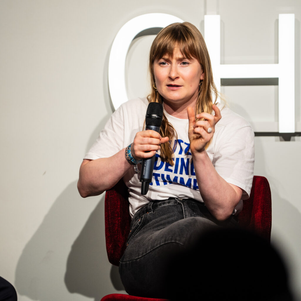 Frau sitzt auf rotem Sessel, hält Mikrofon in der rechten Hand, trägt weißes T-Shirt mit blauem Text und spricht vor weißer Wand mit großem Buchstaben im Hintergrund