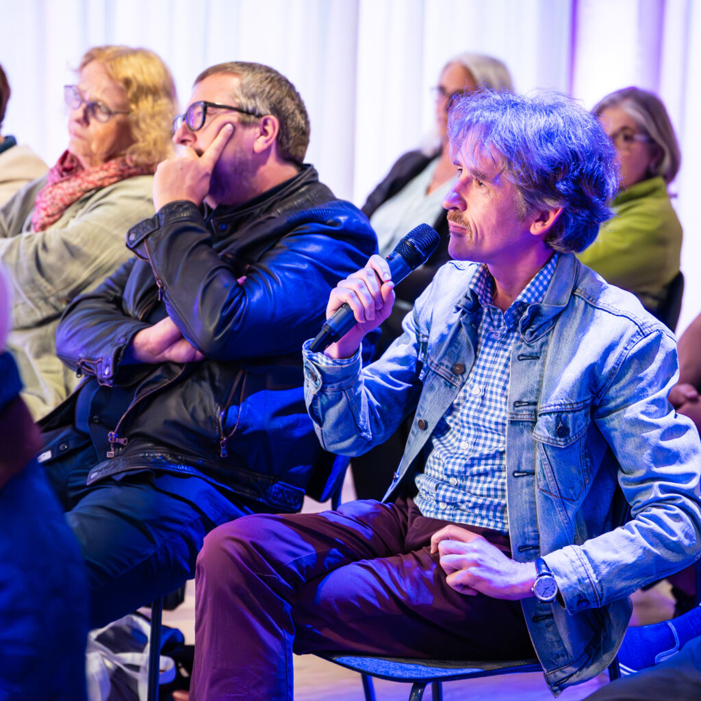 Mann mit Jeansjacke hält Mikrofon und spricht in einer Diskussionsrunde zum Thema Demokratie und Zivilgesellschaft, umgeben von aufmerksam zuhörenden Personen.