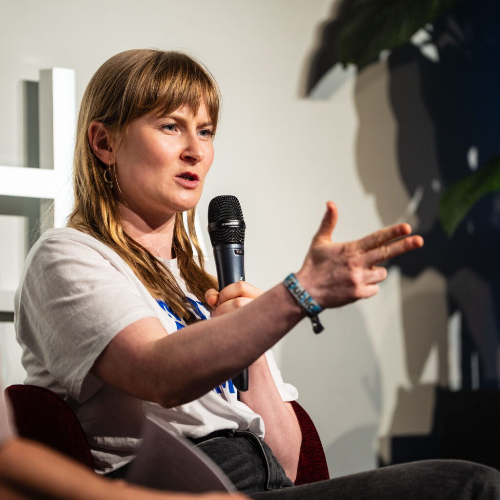 Frau mit mittellangen blonden Haaren hält Mikrofon und gestikuliert mit ausgestreckter Hand während einer Diskussion zum Thema Demokratie und Zivilgesellschaft