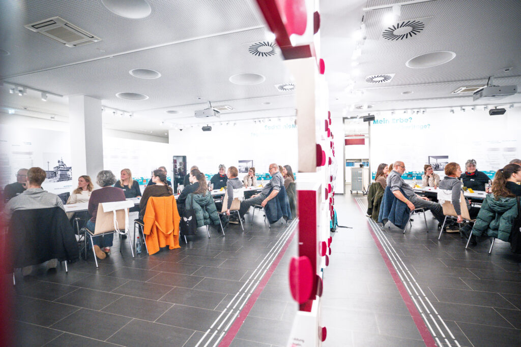 Menschen sitzen an Tischen in einem hellen, modernen Raum mit Spiegelwand und schwarzem Boden