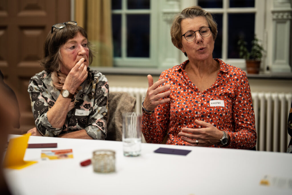 Zwei Frauen sitzen an einem Tisch, eine spricht gestikulierend, die andere hört zu und hält sich die Hand vor den Mund