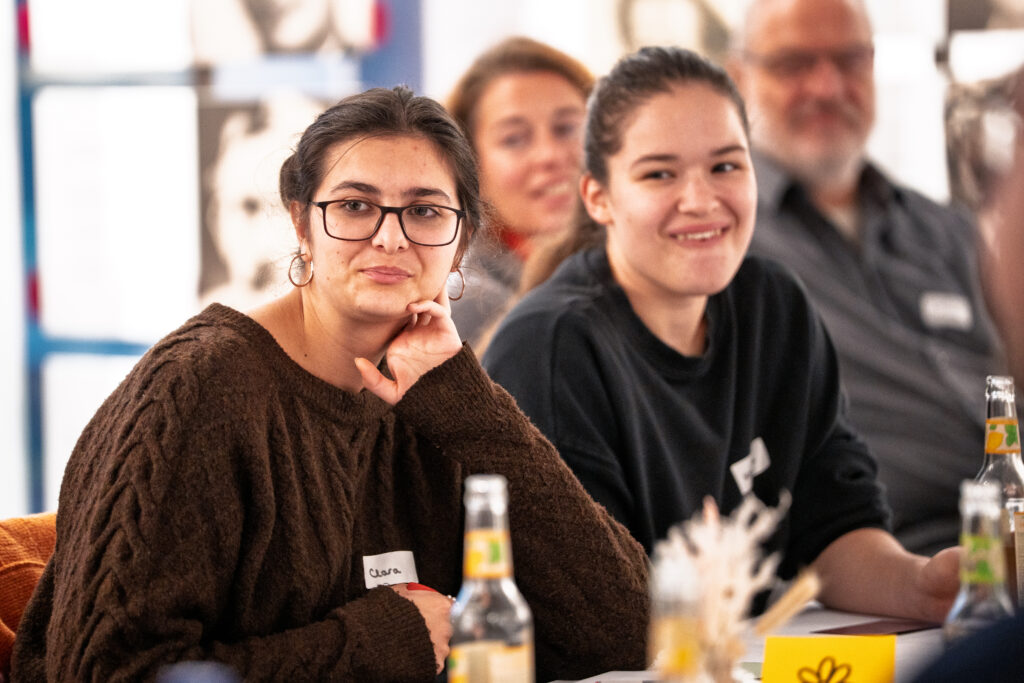 Zwei junge Frauen sitzen nebeneinander an einem Tisch, eine trägt eine Brille und einen braunen Pullover, im Hintergrund zwei weitere Personen unscharf sichtbar