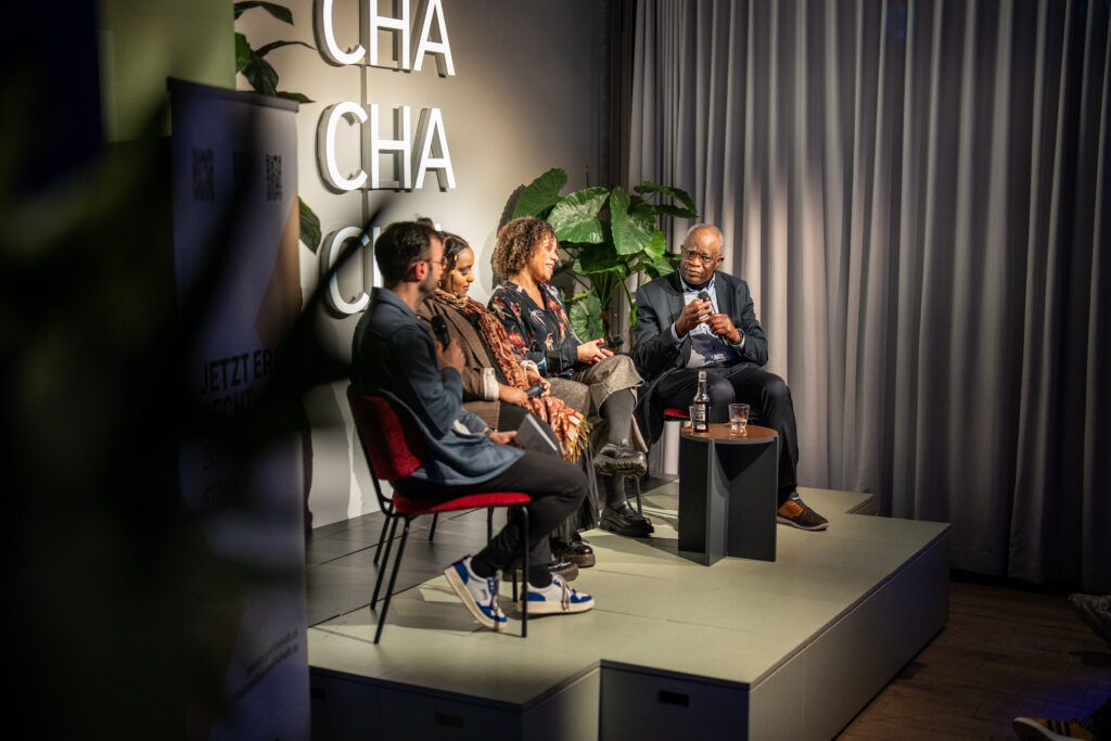 Vier Personen sitzen auf einem Podium, zwei Frauen und zwei Männer, im Gespräch, im Hintergrund große Buchstaben CHA CHA CHA