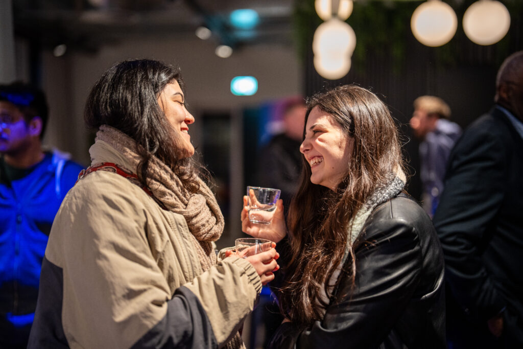 Zwei Frauen im Gespräch, eine hält ein Glas, beide lächeln in einer Innenraumumgebung mit unscharfem Hintergrund und weiteren Personen.