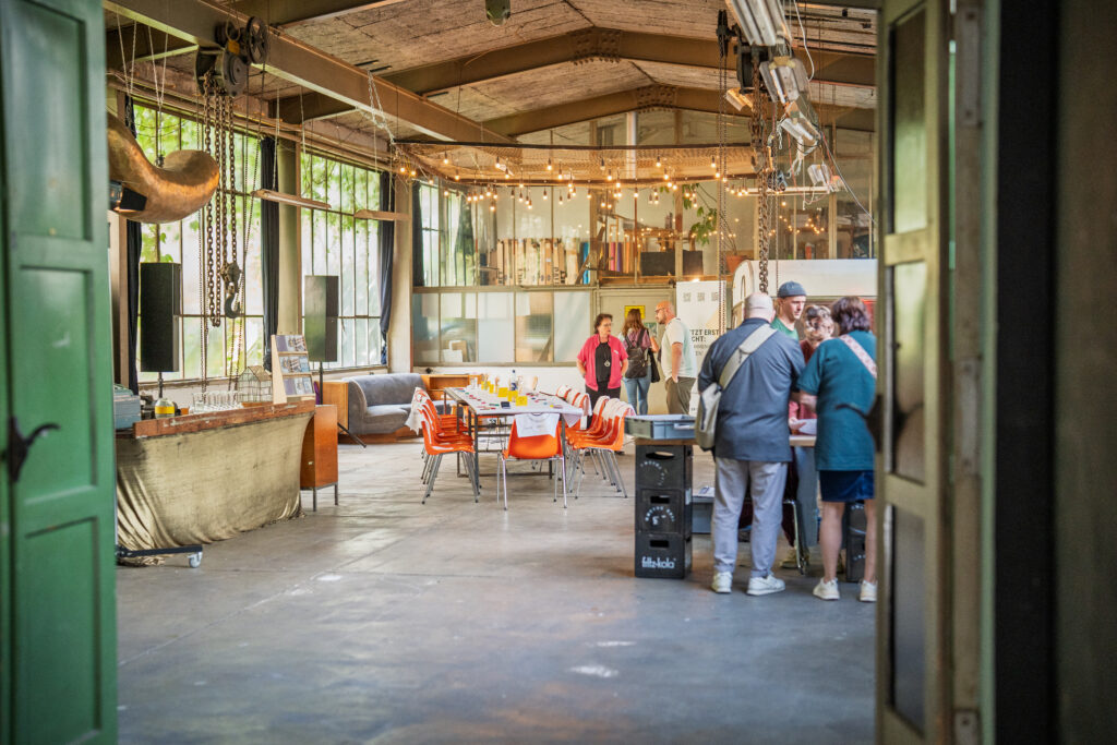 Innenraum einer Industriehalle mit großen Fenstern, Holzbalkendecke, mehreren Menschen, Tischen und Stühlen sowie hängenden Lichterketten