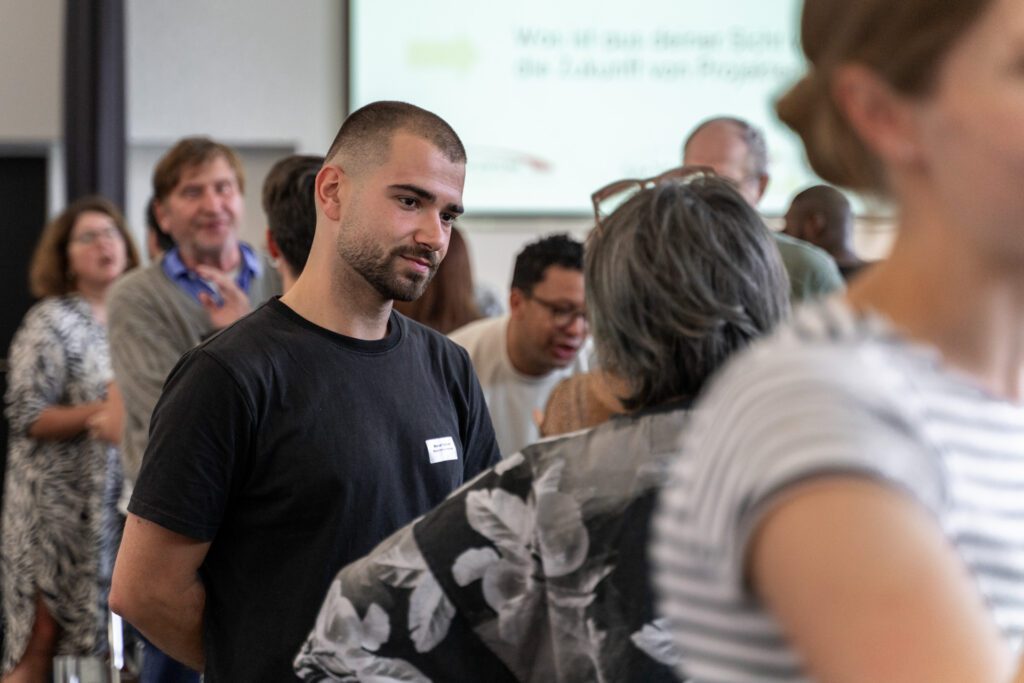 Mann mit kurzem Haar und Bart in schwarzem T-Shirt steht in einer Gruppe von Menschen und spricht mit einer Frau mit grauem, schulterlangem Haar und gemustertem Oberteil