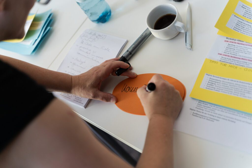 Person schreibt mit schwarzem Stift auf orangefarbenes Oval auf einem Tisch mit Notizblock, Kaffeetasse und Dokumenten
