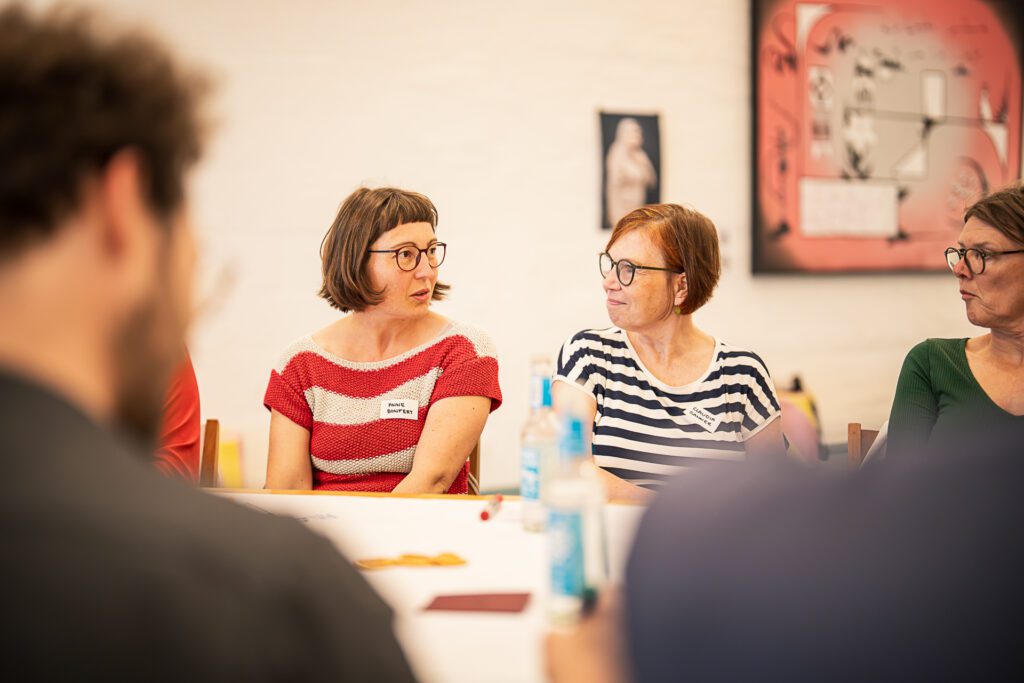 Zwei Frauen mit Brille sitzen an einem Tisch und unterhalten sich, im Vordergrund unscharfe Personen