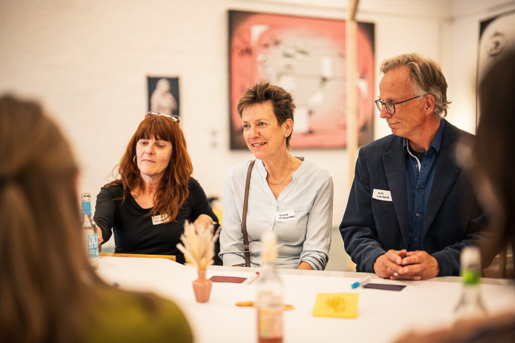 Drei Personen sitzen an einem Tisch, zwei Frauen und ein Mann, alle mit Namensschildern, im Hintergrund ein gerahmtes Bild an der Wand