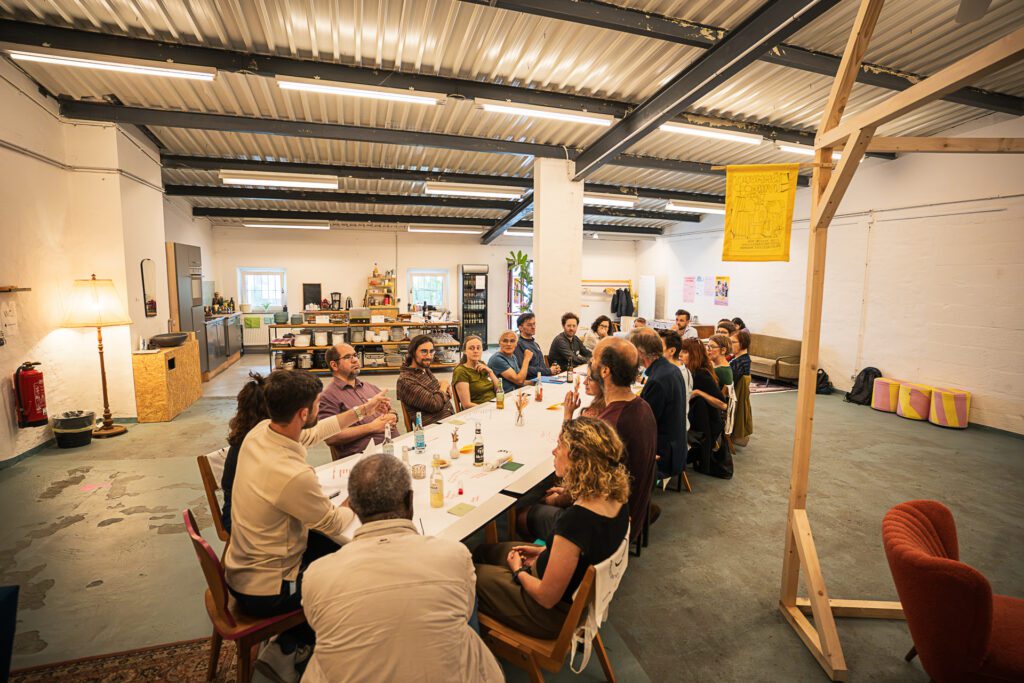 Gruppe von Menschen sitzt an einem langen Tisch in einem hellen, offenen Raum und führt eine Diskussion