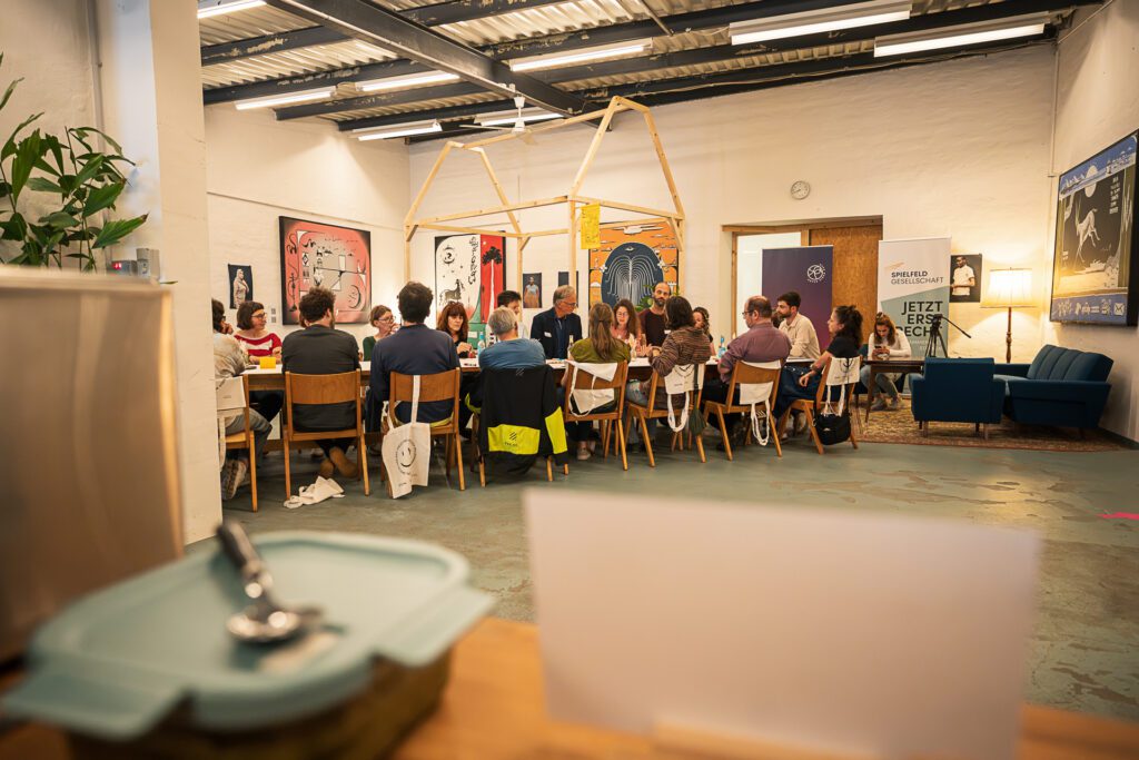 Gruppe von Menschen sitzt in einem Raum in Stuhlkreis und diskutiert, im Hintergrund Poster an den Wänden und eine blaue Couch