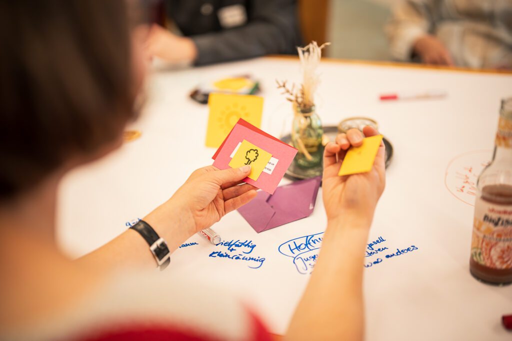 Person hält bunte Karten über einem Tisch mit handschriftlichen Notizen und einer Glasflasche
