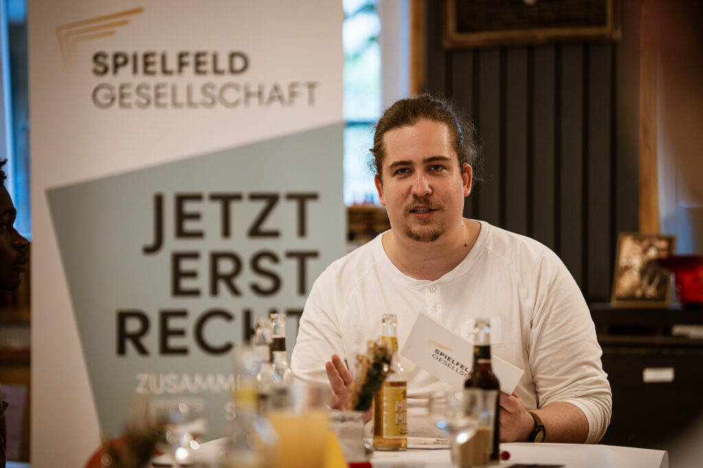 Mann mit weißem Shirt sitzt an einem Tisch mit Getränken und spricht vor einem Banner mit der Aufschrift 'SPIELFELD GESELLSCHAFT JETZT ERST RECHT'.
