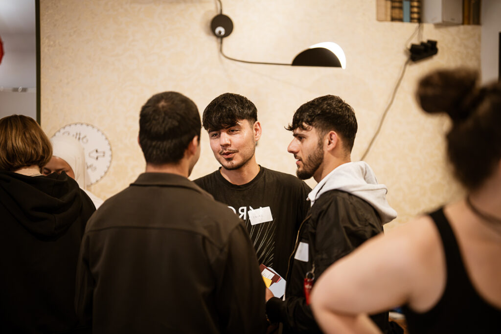 Drei junge Männer im Gespräch in einem Innenraum mit beige gestrichener Wand und runder Lampe an der Wand