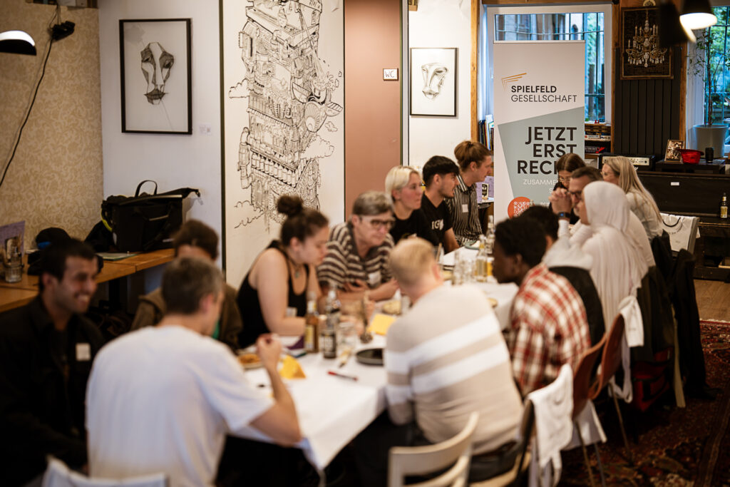 Gruppe von Menschen sitzt an einem langen Tisch in einem Raum mit Kunst an den Wänden und einem Banner mit der Aufschrift 'SPIELFELD GESELLSCHAFT JETZT ERST RECHT'