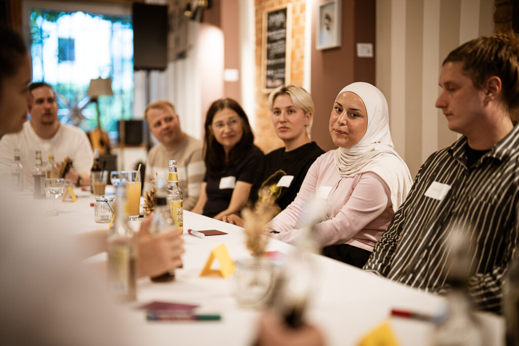 Gruppe von Menschen unterschiedlichen Alters und Geschlechts sitzt an einem langen Tisch in einem hellen Raum und hört aufmerksam zu
